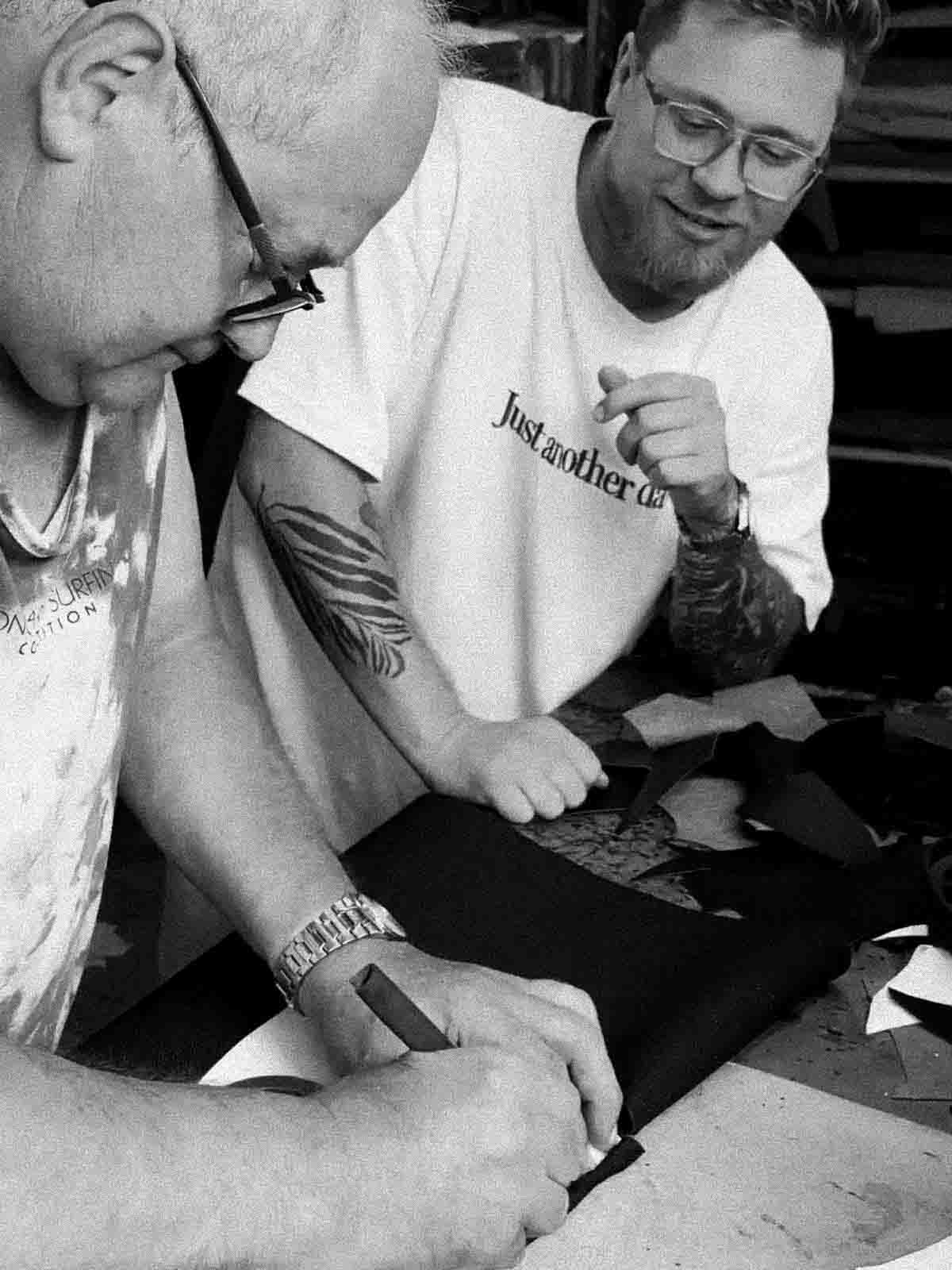Two men working on a project together, one wearing a 'just another' shirt.